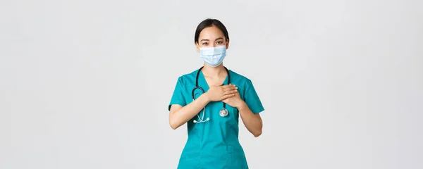 Covid-19, maladie du coronavirus, concept des travailleurs de la santé. Amical bienveillant asiatique femme médecin, médecin en masque médical et gants, tenant la main sur le cœur et souriant, fond blanc — Photo