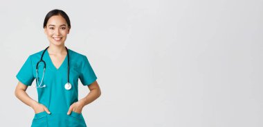 Covid-19, sağlık çalışanları, salgın konsepti. Kendinden emin gülümseyen güzel Asyalı doktor, kararlı ve neşeli görünen doktor, önlük ceplerinde el ele tutuşan, klinikteki hastaları muayene eden.