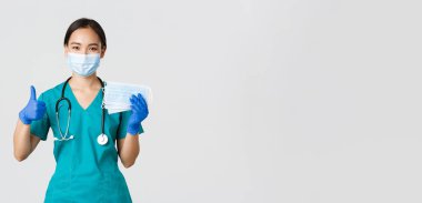 Covid-19, koronavirüs hastalığı, sağlık çalışanları konsepti. Gülümseyen Asyalı doktor, önlüklü hemşire ve lastik eldiven, baş parmak ve tıbbi maskeler, beyaz arka plan