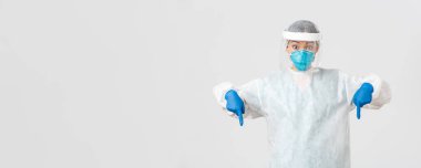 Covid-19, koronavirüs hastalığı, sağlık çalışanları konsepti. Şaşırmış ve heyecanlı kadın doktor, kişisel koruyucu ekipman hekimi parmakları aşağıda, beyaz arka planda duruyor.