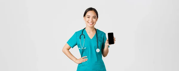 Covid-19, pracownicy służby zdrowia i koncepcja medycyny online. Profesjonalna kobieta azjatycki lekarz, pielęgniarka w fartuchu pokazując ekran smartfona i uśmiechnięty, pokazując aplikację internetową wizytę, białe tło — Zdjęcie stockowe