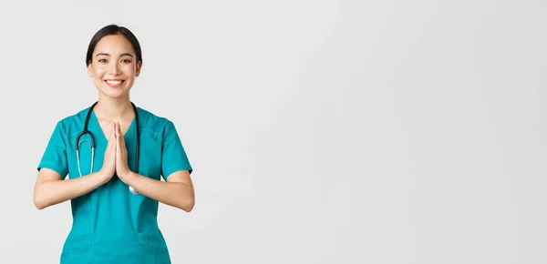 Ковід-19, медичні працівники та запобігання концепції вірусу. Усміхаючись красива азіатська медсестра, лікар в скрабах посміхається, тримає руки разом над грудьми в самовираженні, вітаючи жест — стокове фото