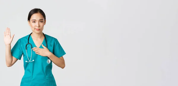 Covid-19, pracownicy służby zdrowia i zapobieganie koncepcji wirusa. Uśmiechnięta, szczera, azjatycka lekarka, stażystka w kitlu, składająca przysięgę, podnosząca jedną rękę i trzymająca się za serce, obiecująca, przysięgająca pacjentowi — Zdjęcie stockowe