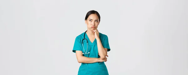 Ковид-19, медицинские работники, пандемия. Раздраженная и обеспокоенная азиатская женщина-врач, медсестра на смене выглядит раздраженной или уставшей, глаза закатываются от скуки и выдоха, стоя на белом фоне в халате — стоковое фото