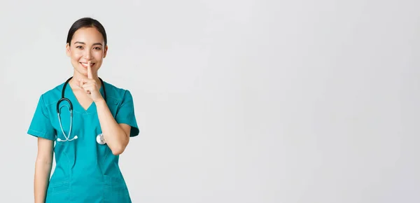 Ковід-19, медичні працівники, пандемія. Весела, усміхнена азіатська медсестра в скрабах має таємницю, робить сюрприз, показуючи чіткий жест, мовчати, стоячи на білому тлі — стокове фото