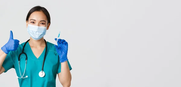 Covid-19, onemocnění koronavirem, koncept zdravotnických pracovníků. Usmívající se krásná asijská lékařka, zdravotní sestra v masce a rukavicích palec nahoru, držte stříkačku s vakcínou, stojící bílé pozadí — Stock fotografie