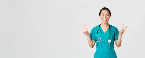 Covid-19, zdravotníci, pandemický koncept. Happy asian female doctor, nurse in scrubs showing peace kawaii gesture and smiling, staying positive during work in hospital, white background — Stock fotografie