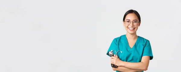 Ковид-19, коронавирусная болезнь, концепция медицинских работников. Уверенная в себе счастливая азиатка-врач, врач в очках, скрещенные руки груди и улыбающаяся, держащая стетоскоп для обследования — стоковое фото