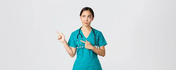 Covid-19, εργαζόμενοι στην υγειονομική περίθαλψη, πανδημία έννοια. Σκεπτικό και αμφίβολο όμορφο ασιατικό γιατρό, νοσοκόμα σε scrubs χαμόγελο, δείχνοντας πάνω αριστερή γωνία, κοιτάζοντας με απρόθυμα διστακτικό πρόσωπο, λευκό φόντο — Φωτογραφία Αρχείου