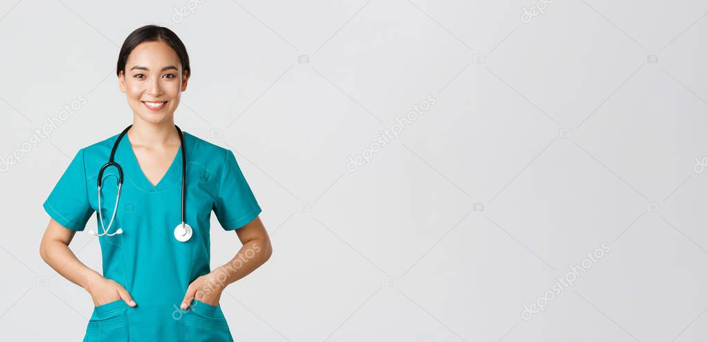 Covid-19, healthcare workers, pandemic concept. Confident smiling pretty asian female doctor, physician looking determined and upbeat, holding hands in pockets of scrubs, examine patients in clinic