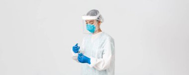 Covid-19, koronavirüs hastalığı, sağlık çalışanları konsepti. Ciddi görünümlü kadın hekim, kişisel koruma ekipmanlarında Asyalı doktor aşıyla birlikte ampul içine şırınga yerleştirin.