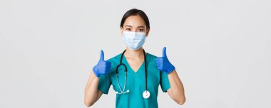 Covid-19, koronavirüs hastalığı, sağlık çalışanları konsepti. Kendinden emin gülümseyen Asyalı doktor, tıbbi maske takan hemşire ve lastik eldiven, onaylama için onay ver, hizmet et, bilgi topla.