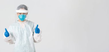 Covid-19, koronavirüs hastalığı, sağlık çalışanları konsepti. Ciddi görünüşlü, kendine güvenen genç Asyalı doktor, kişisel koruma ekipmanlarında enfeksiyoncu baş parmağını kaldırarak tehlikeli bölgeye giriyor.
