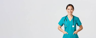 Covid-19, sağlık çalışanları, salgın konsepti. Kendinden emin gülümseyen güzel Asyalı doktor, kararlı ve neşeli görünen doktor, önlük ceplerinde el ele tutuşan, klinikteki hastaları muayene eden.