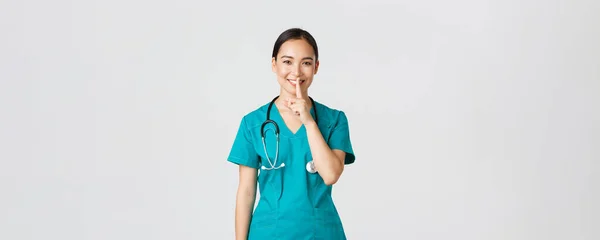 Covid-19, pracownicy służby zdrowia, koncepcja pandemii. Wesoły, uśmiechnięta kobieta azjatycka pielęgniarka w fartuchu mając sekret, co niespodzianka, pokazując milczący gest, shhh siedzieć cicho, stojąc białe tło — Zdjęcie stockowe