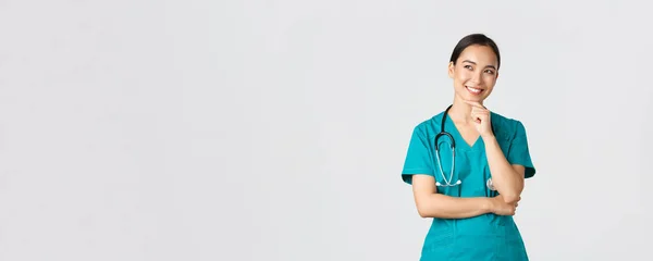Covid-19, zdravotníci, pandemický koncept. Usmívající se potěšen atraktivní asijské žena lékař v křoví, při pohledu v levém horním rohu a myšlení, mají nápad, stojící zamyšlené bílé pozadí — Stock fotografie