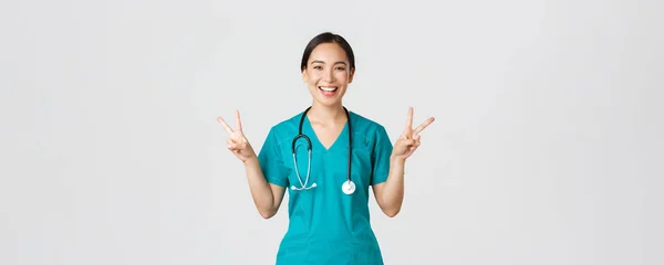 Covid-19, zdravotníci, pandemický koncept. Happy asian female doctor, nurse in scrubs showing peace kawaii gesture and smiling, staying positive during work in hospital, white background — Stock fotografie