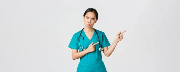 Covid-19, pracownicy służby zdrowia, koncepcja pandemii. rozczarowany i zły azjatycki kobieta terapeuta pielęgniarka w fartuch patrząc zdenerwowany na aparat, dąsając się i wskazując prawym górnym rogu, czekać wyjaśnienie — Zdjęcie stockowe