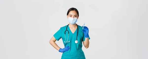 Covid-19, choroba koronawirusowa, koncepcja pracowników służby zdrowia. Uśmiechnięta, pewna siebie azjatycka pielęgniarka w fartuchu i masce medycznej, trzymająca strzykawkę ze szczepionką, wykonująca szczepienia, białe tło — Zdjęcie stockowe