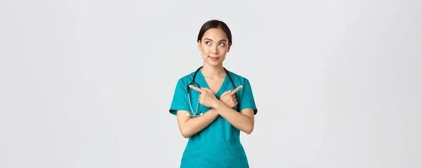 Covid-19, healthcare workers, pandemic concept. Creative and thoughtful pretty asian female nurse, intern have idea, making choice, pointing fingers sideways but looking left, thinking — Stock Photo, Image