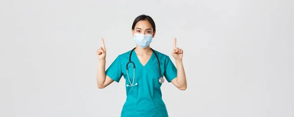 Covid-19, koronavírus betegség, egészségügyi dolgozók koncepciója. Fiatal professzionális ázsiai női orvos, ápolónő orvosi maszkban és köpenyben, ujjal felfelé mutogatva, utat mutatva, reklám — Stock Fotó