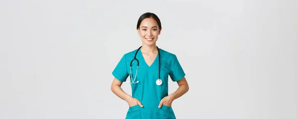 Covid-19, healthcare workers, pandemic concept. Confident smiling pretty asian female doctor, physician looking determined and upbeat, holding hands in pockets of scrubs, examine patients in clinic — Stock Photo, Image