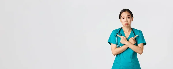 Covid-19, egészségügyi dolgozók, világjárvány. Határozatlan és tudatlan ázsiai női nővér, gyakornok nem tudom, mit válasszon, ujjal oldalra mutat, és vonagló tudatlan, fehér háttér — Stock Fotó