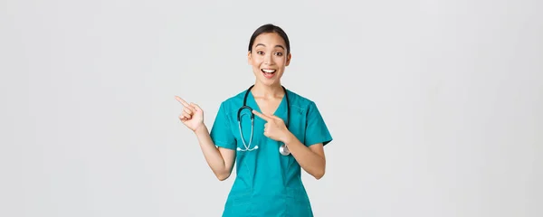 Covid-19, Gesundheitspersonal, Pandemiekonzept. Aufgeregt asiatische Krankenschwester, Arzt in Peelings und Stethoskop, Zeigefinger oben links Ecke. Arzt lächelt erstaunt und zeigt ehrfürchtige Ansage — Stockfoto