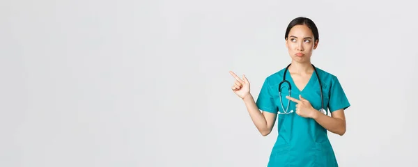 Ковид-19, медицинские работники, пандемия. Разочарованная мрачная, угрюмая азиатская медсестра в халате, недовольная женщина-врач, указывающая на левый верхний угол и хмурящаяся осуждающая — стоковое фото