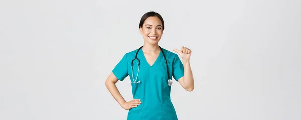 Covid-19, healthcare workers, pandemic concept. Professional confident female asian doctor, nurse in scrubs pointing at herself and smiling, guarantee she knows what do, white background — Stock Photo, Image