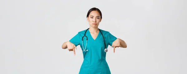 Covid-19, pracownicy służby zdrowia, koncepcja pandemii. Zdenerwowana i rozczarowana dąsająca się azjatycka terapeutka, pielęgniarka w fartuchu wskazująca palcem w dół, dąsająca się smutna, pokazująca coś z żalem — Zdjęcie stockowe