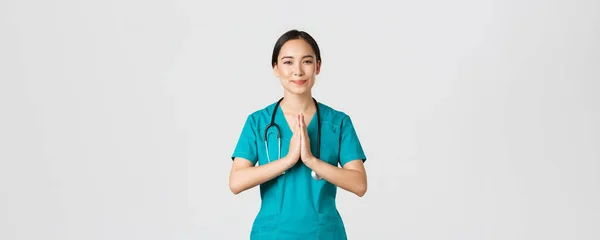 Covid-19, pracownicy służby zdrowia i zapobieganie koncepcji wirusa. Uśmiechnięta i opiekuńcza, ładna azjatycka lekarka, lekarka w fartuchu, wykazująca namaste, modlący się gest, białe tło — Zdjęcie stockowe