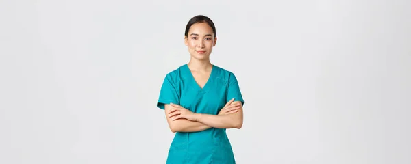 Covid-19, sjukvårdspersonal, pandemikoncept. Självsäker leende asiatisk sjuksköterska i skrubbar stående självsäker, kors armar bröstet. Professionell läkare redo för operation, stående vit bakgrund — Stockfoto