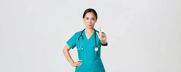 Covid-19, egészségügyi dolgozók, világjárvány. Komoly kinézetű, hivatásos női ázsiai orvos köpenyben, orvos remegő ujj tilalomban, figyelmeztető gesztus, megtilt valamit. — Stock Fotó