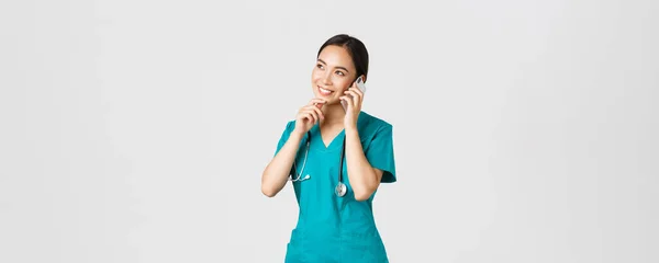 Covid-19, healthcare workers and preventing virus concept. Portrait of smiling asian female doctor, intern in scrubs talking on phone and looking thoughtful, thinking or making choice — Stock Photo, Image