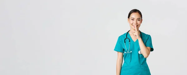 Covid-19, egészségügyi dolgozók, világjárvány. Vidám, mosolygó ázsiai nővér köpenyben, amelynek titkát, hogy meglepetés, mutatja shush gesztus, shhh csendben, álló fehér háttér — Stock Fotó