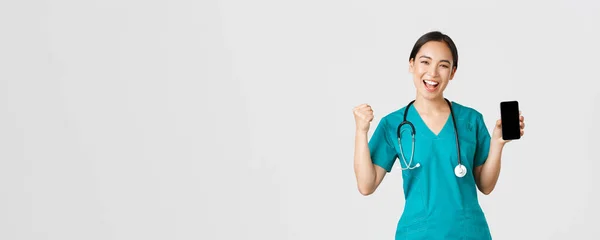 Covid-19, lucrătorii din domeniul sănătății și conceptul de medicină online. Succes vesel asiatic femeie doctor, asistenta medicala in scrubs pumnul pompa în bucurie, arătând aplicația ecran smartphone și în căutarea mulțumit la aparat de fotografiat — Fotografie, imagine de stoc