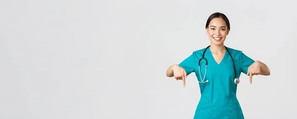 Ковід-19, медичні працівники, пандемія. Усміхнена приємна азіатська жінка-лікар, терапевт або лікар в скрабах зі стетоскопом, вказує пальці вниз, показує клінічний банер — стокове фото