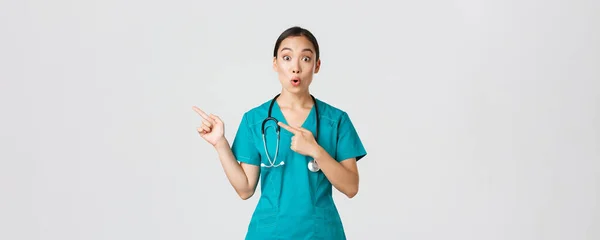 Covid-19, healthcare workers, pandemic concept. Impressed and amazed female asian nurse in scrubs asking question with intrigued expression. Doctor looking thrilled and pointing upper left corner — Stock Photo, Image