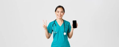 Covid-19, sağlık çalışanları ve online tıp konsepti. Gülümseyen kendinden emin Asyalı kadın doktor, önlüklü doktor barış işareti ve mobil ekranı gösteriyor, danışma uygulamasını destekliyor, beyaz arka plan