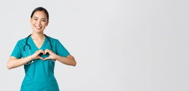 Covid-19, sağlık çalışanları, salgın konsepti. Sevecen Asyalı doktor, önlüklü kadın hemşire kalp hareketi ve gülümseme gösteriyor, beyaz geçmişi olan hastalarla ilgileniyor.