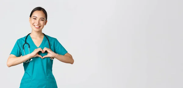 Covid-19, sağlık çalışanları, salgın konsepti. Sevecen Asyalı doktor, önlüklü kadın hemşire kalp hareketi ve gülümseme gösteriyor, beyaz geçmişi olan hastalarla ilgileniyor.
