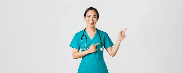 Covid-19, trabajadores de la salud, concepto pandémico. Sonriente alegre asiática médico, terapeuta en matorrales señalando los dedos esquina superior derecha, invitando a chequear, recomendar vacunar — Foto de Stock