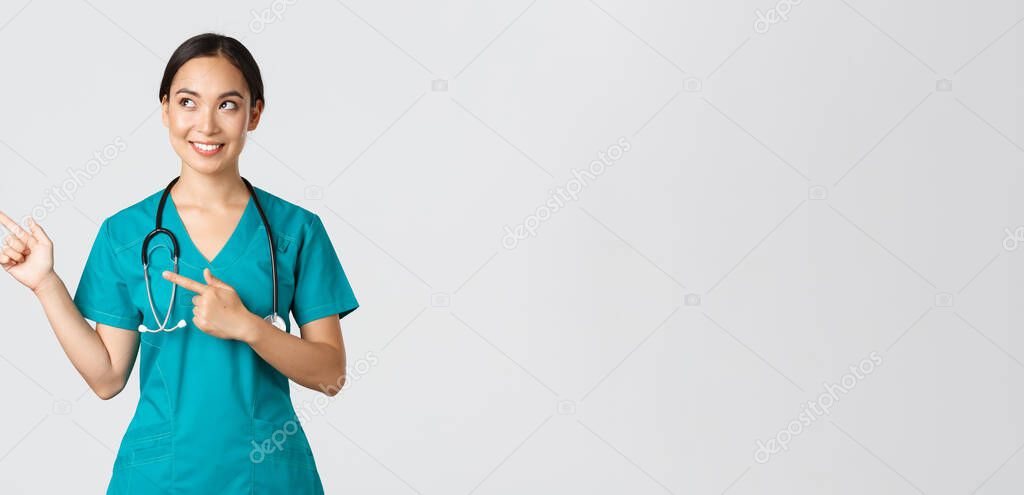 Covid-19, healthcare workers, pandemic concept. Intrigued beautiful asian female intern, nurse in scrubs looking and pointing upper left corner with curious dreamy smile, white background