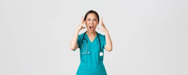 Covid-19, healthcare workers and preventing virus concept. Annoyed and angry, pissed-off asian doctor, female nurse in scrubs going mad, screaming in rage, shaking hands outraged, white background