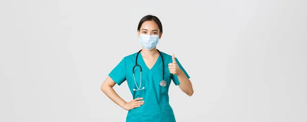 Covid-19, coronavirus disease, healthcare workers concept.自信地微笑着的亚裔医生、女护士或戴着医疗面罩和擦拭的医生，显示出大拇指向上能保证质量、礼貌 — 图库照片