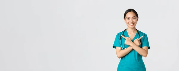 Covid-19, healthcare workers, pandemic concept. Smiling beautiful asian nurse, intern or doctor in scrubs pointing sideways and looking at camera, showing variants, two choices, white background — Stock Photo, Image