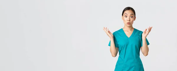 Ковид-19, медицинские работники, пандемия. Удивлённая и изумленная азиатская медсестра в халате, поднимающая руки вверх, выглядящая испуганной, узнавшая большие новости, больница получила коронавирусную вакцину, белый фон — стоковое фото