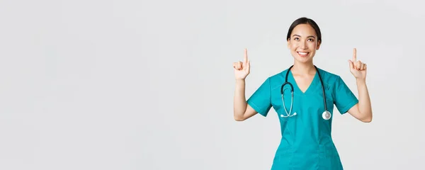 Ковід-19, медичні працівники, пандемія. Усміхнена красива азіатська медсестра, лікар в скрабах вказуючи і дивлячись вгору з задоволеною усмішкою, щаслива, щоб показати чудову промо-пропозицію, білий фон — стокове фото
