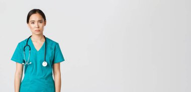 Covid-19, sağlık çalışanları, salgın konsepti. Bitkin düşmüş genç Asyalı kadın hemşire, hastanede vardiyadan sonra yorgun görünen doktor, yorgunluktan üzgün görünüyor, önlüklü beyaz arka plan